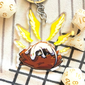 May include: A white dragon with yellow wings is curled up on a brown log. The dragon is a keychain charm with a silver key ring.