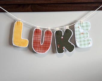 Könnte beinhalten: Ein Stoffbuchstaben-Banner, das den Namen LUKE buchstabiert und an einer weißen Schnur hängt. Der Buchstabe L ist gelb mit Sternmuster, U ist rot kariert, K ist dunkelgrün mit kleinen Pilzmotiven und E ist hellgrün kariert mit weißen Enten.
