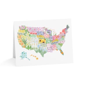 May include: A white greeting card featuring a watercolor illustration of the United States map filled with colorful flowers. The flowers are arranged to represent different states. Additional floral elements are present below the map.