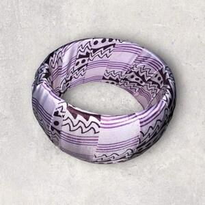 May include: A purple and white fabric bangle bracelet with a geometric pattern.