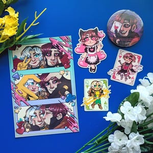 May include: A collection of anime-style merchandise featuring a rectangular photo strip with three panels, a circular pinback button, and two rectangular stickers. The artwork depicts characters with pink and white hair, hearts, and playing card motifs.