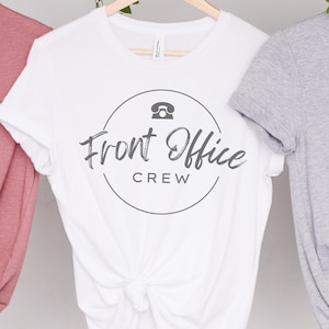 Peut inclure: T-shirt blanc avec un graphique circulaire gris qui dit "Front Office Crew" et une icône de téléphone.
