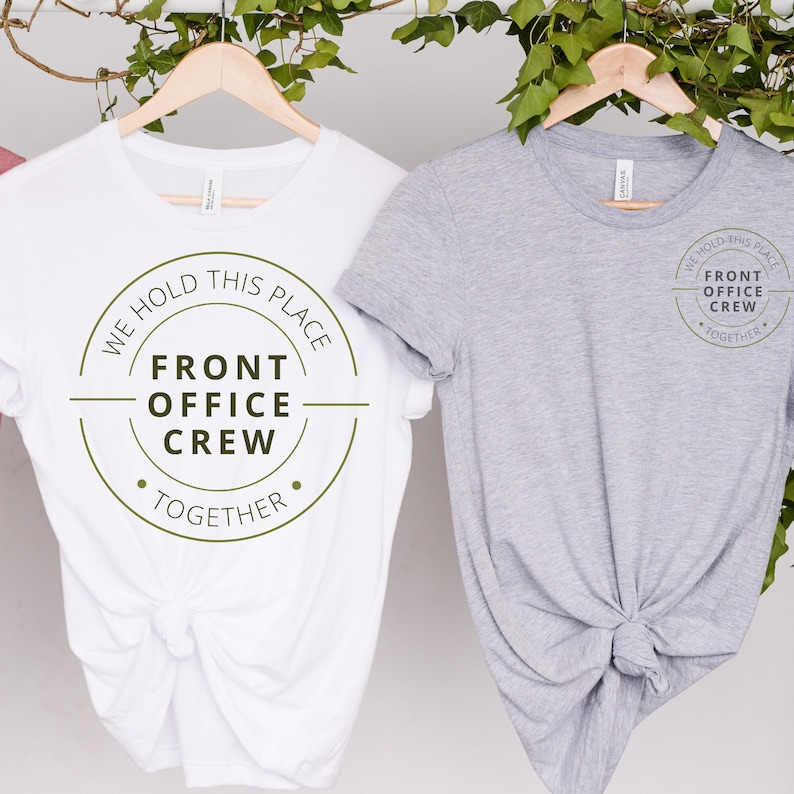 K&ouml;nnte beinhalten: Zwei wei&szlig;e T-Shirts mit einem gr&uuml;nen Kreis-Motiv, auf dem "We hold this place, Front Office Crew, Together" steht. Ein Shirt ist wei&szlig; und das andere grau.