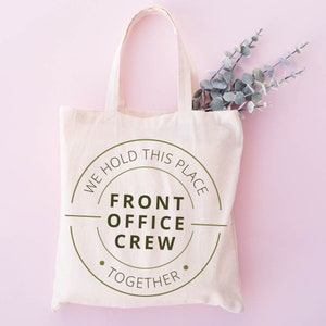 K&ouml;nnte beinhalten: Eine wei&szlig;e Canvas-Tragetasche mit einem gr&uuml;nen Kreislogo, auf dem "We hold this place together" und "Front Office Crew" in der Mitte steht.
