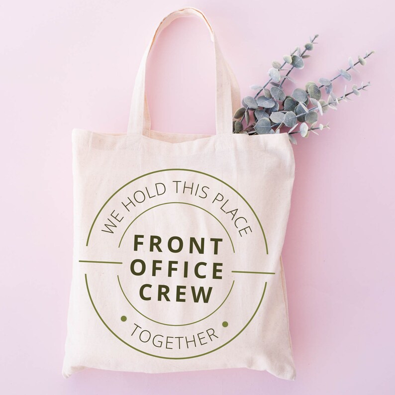 K&ouml;nnte beinhalten: Eine wei&szlig;e Canvas-Tragetasche mit einem gr&uuml;nen Kreislogo, auf dem "We hold this place together" und "Front Office Crew" in der Mitte steht.