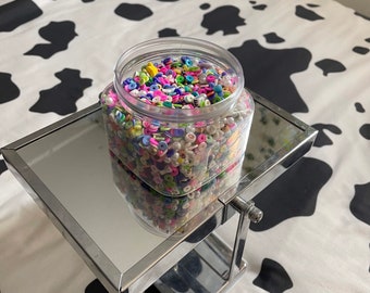 May include: A clear plastic jar filled with colorful letter beads. The jar is sitting on a silver metal stand with a mirrored surface. The stand is on a white and black cow print surface.