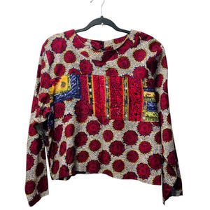May include: A long-sleeve shirt featuring a repeating pattern of dark red circular motifs on a white background, with a central rectangular panel displaying vertical stripes in red, yellow, blue, and black, hanging on a black plastic hanger.
