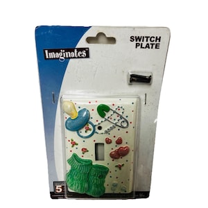 May include: A white switch plate in original packaging labeled Imaginates, featuring a raised blue pacifier, a green safety pin with the word BABY, and green baby booties. The plate is decorated with small red and blue dots and pink rose accents.