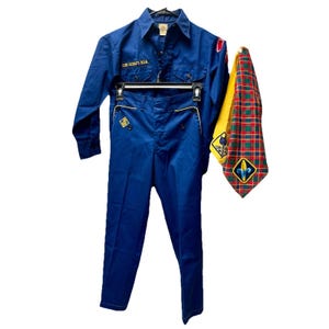 May include: A blue Cub Scouts BSA uniform set including a long-sleeved button-down shirt with embroidered patches, matching blue trousers with yellow piping, and a yellow, red, and green plaid neckerchief featuring a yellow fleur-de-lis patch.
