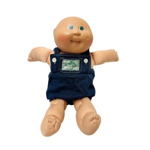 May include: A bald doll with a round, light-toned vinyl head, rosy cheeks, and green eyes. The doll has a soft fabric body and is wearing dark blue denim overalls with white buttons and a small green embroidered patch on the chest pocket.