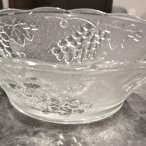 May include: A clear glass bowl featuring a textured surface with embossed grape clusters and vine leaf patterns. The bowl has a scalloped, wavy rim and a circular base, showcasing a transparent design that catches light across its detailed exterior.
