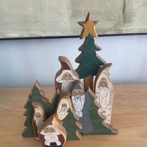 May include: A set of wooden Christmas ornaments featuring Santa Claus figures and pine trees. The ornaments are painted in various shades of green, red, white, and brown. The figures have whimsical faces and are dressed in traditional Santa Claus attire.
