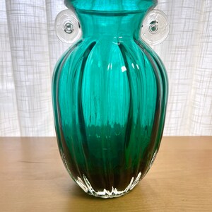 May include: A teal green glass vase with clear glass handles. The vase has a ribbed design and a rounded bottom.