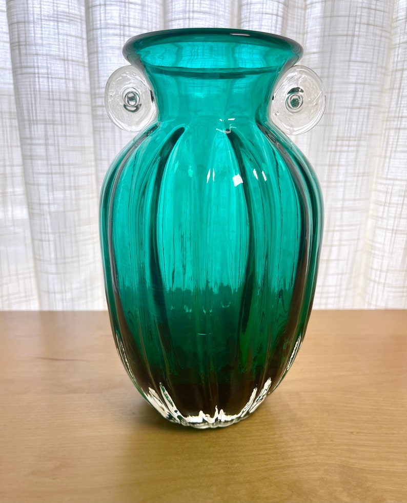 May include: A teal green glass vase with clear glass handles. The vase has a ribbed design and a rounded bottom.