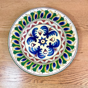 May include: A round ceramic plate with a colourful floral design. The plate features a central motif of blue and yellow flowers with a star in the centre, surrounded by a border of green and yellow leaves.