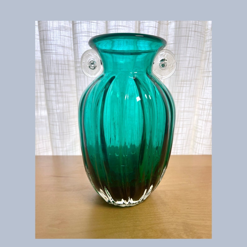May include: A teal green glass vase with white glass handles. The vase has a ribbed design and a rounded body.