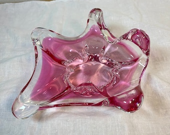 May include: A pink glass bowl with a unique, organic shape. The bowl has a clear glass base and a pink glass top. The bowl is perfect for holding jewelry, trinkets, or other small items.