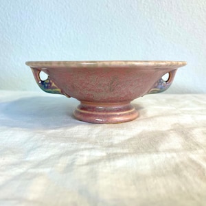 Può includere: Una piccola ciotola in ceramica rosa con due manici. La ciotola ha una superficie testurizzata e una base rialzata. I manici sono decorati con un motivo floreale in tonalità blu e verdi.