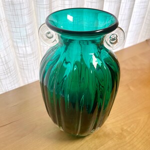 May include: A teal green glass vase with clear glass handles. The vase has a ribbed design and a rounded bottom.