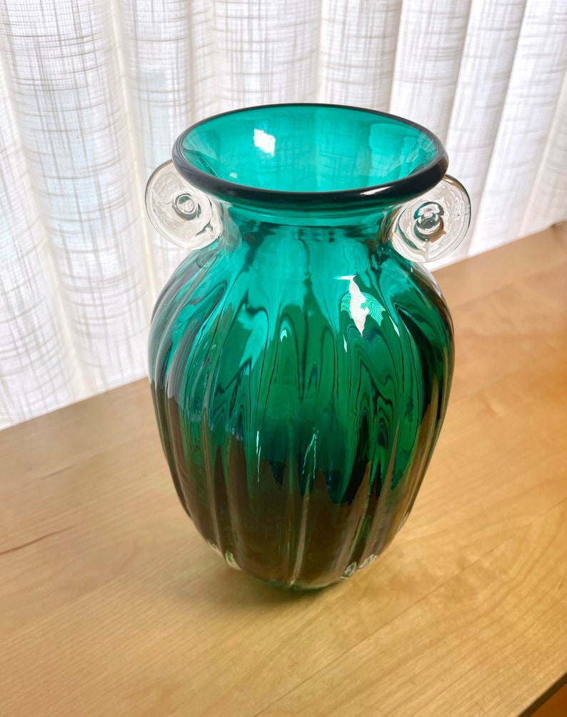 May include: A teal green glass vase with clear glass handles. The vase has a ribbed design and a rounded bottom.