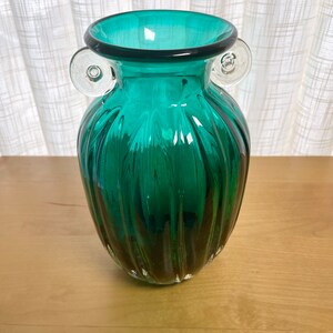 May include: A teal green glass vase with clear glass handles. The vase has a ribbed design and a rounded body.