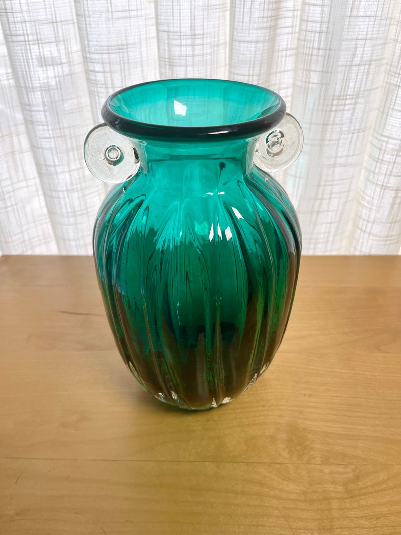 May include: A teal green glass vase with clear glass handles. The vase has a ribbed design and a rounded body.