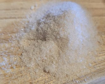 May include: A pile of coarse, light-colored granules, possibly sugar or salt, sits on a light-brown wooden surface. The granules are clustered together, creating a small mound with a slightly uneven texture. The lighting highlights the crystalline structure.
