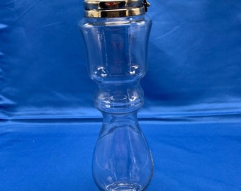 May include: A clear glass bottle with a silver lid and a hinged closure. The bottle has a wide base and a narrow neck. The bottle is empty.
