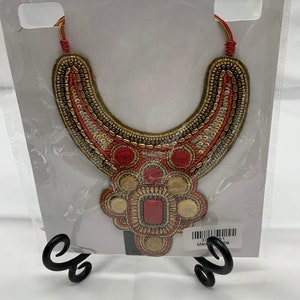 May include: A red and gold beaded necklace with a large, ornate pendant. The pendant is made of red and gold beads and has a square-shaped stone in the center. The necklace is displayed on a white background with a black stand.