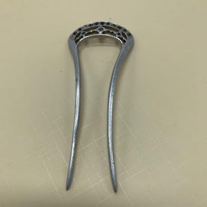 May include: A silver-colored hair pin with a decorative, openwork design at the top. The pin has two long, slender prongs. The top section features an intricate pattern of small shapes and curves.