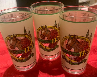 Può includere: Tre bicchieri di vetro trasparente con sfondo bianco e dettagli in verde e rosso. Ogni bicchiere presenta un'illustrazione di un cavallo e un fantino con il testo "Churchill Downs Louisville, Kentucky" e "Kentucky Derby 113". I bicchieri sono su una superficie rossa.