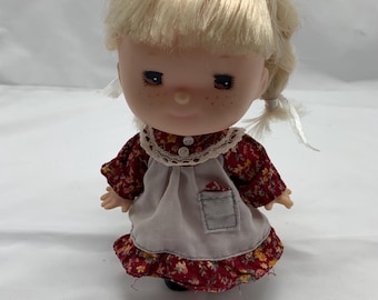May include: A vintage doll with blonde hair, freckles, and closed eyes. The doll wears a red floral dress with a white apron and a lace collar. The doll has a small pocket on the apron and is wearing a black headband.