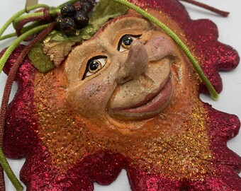 May include: A red and gold glitter ornament shaped like a face with a large nose and a wide smile. The ornament has a green felt hat with black berries on top.