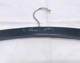 May include: A black wooden clothes hanger featuring a silver metal hook at the top. The front of the hanger is inscribed with the white cursive text "i love you" centred along the horizontal bar, providing a clear and legible message.