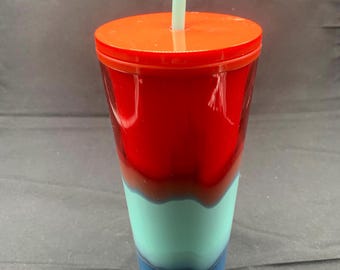 May include: A multi-coloured tumbler with a red lid and a pale green straw. The body of the cup features a gradient of red, teal, and blue. This reusable cup is suitable for cold drinks.
