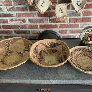 May include: Three woven baskets with intricate geometric patterns in shades of tan and brown. The baskets are oval and round, displayed on a gray surface. A snow globe and a banner with letters are in the background.