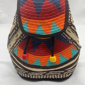 May include: A colorful woven backpack with a geometric design. The flap features a vibrant pattern of red, orange, blue, and yellow. The bag has a drawstring closure with red and yellow bead accents. The sides are woven with a natural fiber.