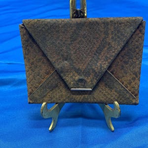 May include: A small, brown envelope-shaped purse with a textured surface and a metal clasp. The purse is displayed on a miniature gold easel against a blue background. The purse has a unique, folded design.