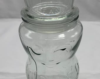May include: Clear glass peanut jar with a top hat-shaped lid. The jar is shaped like a cartoon character with a smiling face and the word "PEANUT" is embossed on the lid. The jar has a wide base and a narrow neck.