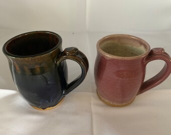 May include: Two handmade ceramic mugs, one with a dark blue glaze and brown accents, and the other with a light pink glaze. Both mugs have a rounded shape and a handle.