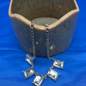 May include: Silver-toned necklace featuring rectangular clear crystals. The necklace has a chain design and is displayed against a blue background. The crystals are set in a silver metal. The necklace is near a brown ceramic bowl.