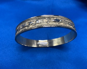 May include: A silver bangle bracelet with a decorative design of interlocking circles and leaves. The bracelet is smooth and shiny.