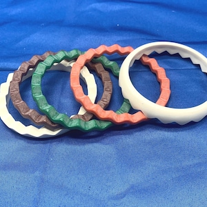 May include: A collection of colorful, wavy-edged bangles in white, brown, green, and coral. The bracelets are arranged on a blue surface, showcasing their unique, textured design. Each bangle has a diameter of approximately 3 inches.