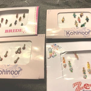 May include: Four boxes of decorative bindi stickers. The boxes are pink and white with clear windows displaying the bindis. The bindis are various colors and designs, including floral and jewel-like patterns. One box is labeled "BRIDE."