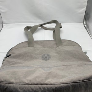 May include: A gray tote bag with a zipper closure and two top handles. The bag has a small circular logo with the text "Kipling" and a star inside.