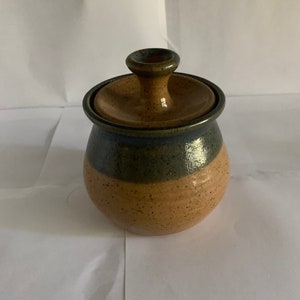 May include: A handcrafted ceramic jar with a lid. The jar is round with a speckled tan base, a dark blue-green band, and a brown lid with a small handle. The pottery has a rustic, earthy aesthetic.