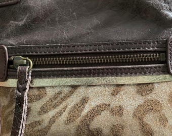 May include: Close-up of a brown leather handbag with a zip closure. The bag features a section with a tan and brown animal print pattern. The leather has a distressed appearance, and the hardware is brass-coloured.