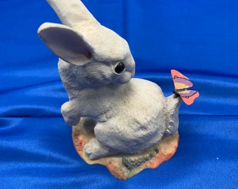 May include: White ceramic rabbit figurine with a pink and purple butterfly on its tail. The rabbit is sitting on a textured base.