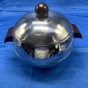 May include: A shiny, silver-colored serving dish with a lid and two wooden handles. The lid has a dark brown, round knob. The dish has a decorative penguin design around the base. The dish is sitting on a blue surface.