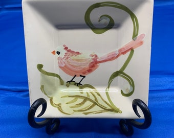 May include: A white ceramic square dish with a green vine and a pink bird painted on it. The dish is on a black stand.